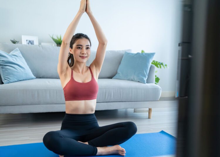 Chica joven y atractiva asiatica en ropa deportiva se siente feliz en la sala de estar de la casa practicando yoga o pilates, realizando entrenamiento en casa.